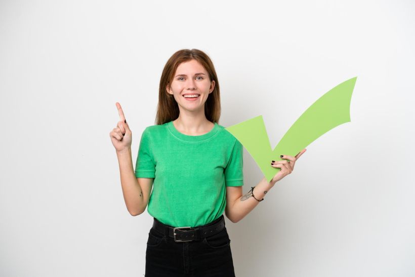 Young woman isolated on white background holding a check icon and pointing up.
