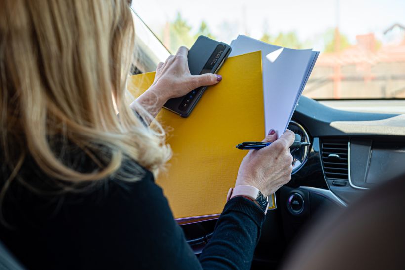 Mujer de negocios rubia, de mediana edad, con gafas, trabajando con documentos en el coche, vista de espaldas.