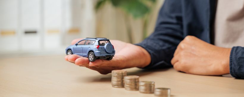 Primer plano de la mano de un hombre sosteniendo un coche de juguete azul con pilas de monedas apiladas cerca, destacando los pagos de un pr&eacute;stamo para la financiaci&oacute;n de un autom&oacute;vil.