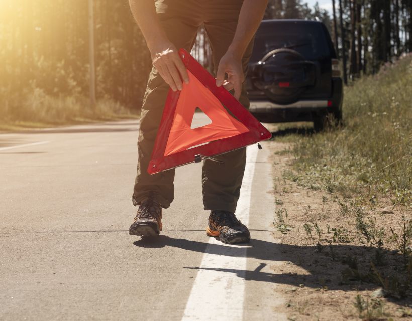 Primer plano de unas manos masculinas colocando una se&ntilde;al de precauci&oacute;n con un tri&aacute;ngulo rojo en el arc&eacute;n de una carretera, cerca de un coche averiado.