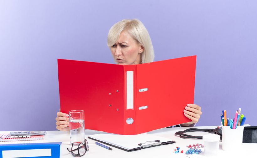 Mujer adulta despistada sentada en un escritorio con herramientas de oficina, sosteniendo y mirando una carpeta de archivos aislada en una pared morada.