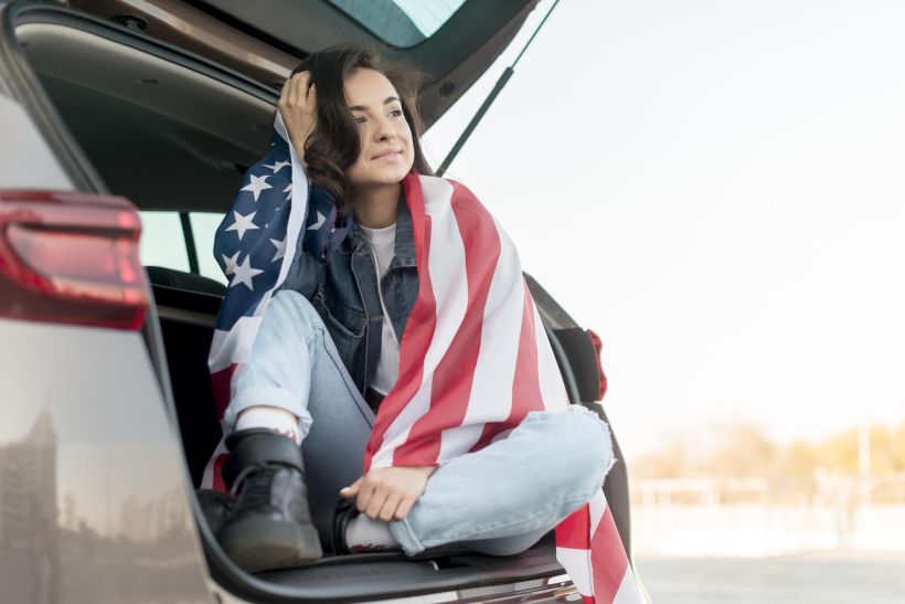 Una joven sostiene una gran bandera estadounidense en el maletero de su coche.