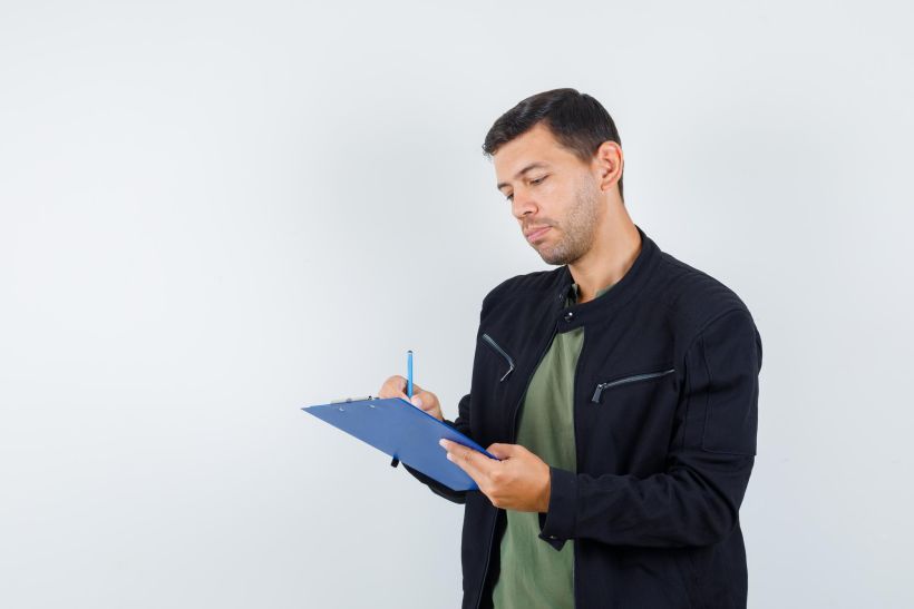Joven var&oacute;n tomando notas en un portapapeles, vestido con camiseta y chaqueta, y con aspecto ocupado.