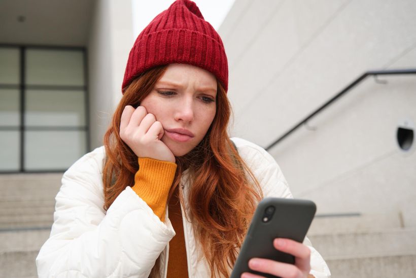 Retrato de una chica pelirroja triste que parece molesta y decepcionada mirando la pantalla de su smartphone.