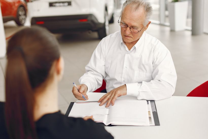Anciano sentado en el sal&oacute;n de un concesionario de coches hablando con el gerente.