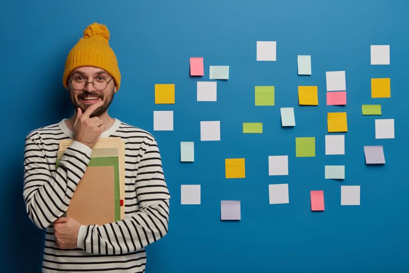 Un alegre estudiante sostiene la barbilla con las manos, con un cuaderno y un libro de texto, y utiliza notas adhesivas en una pared azul.